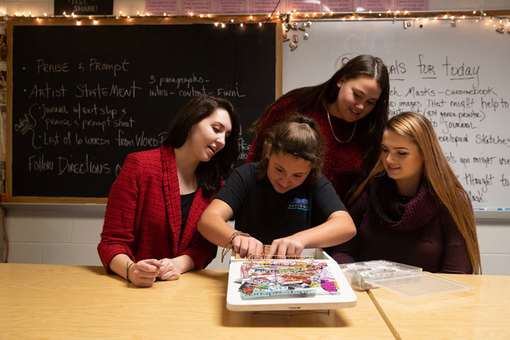 Three female students watch a fourth create a page of the co-constructed book.