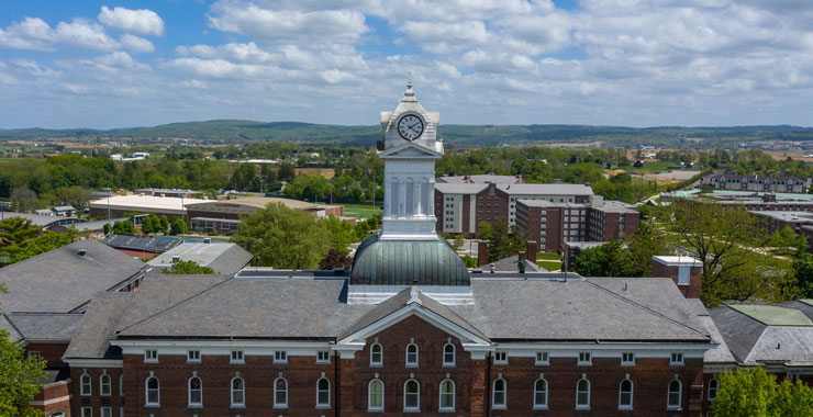 Aerial view of front of Old Main, clock tower centered, with view of South Campus and cloudy, blue skies in the background.
