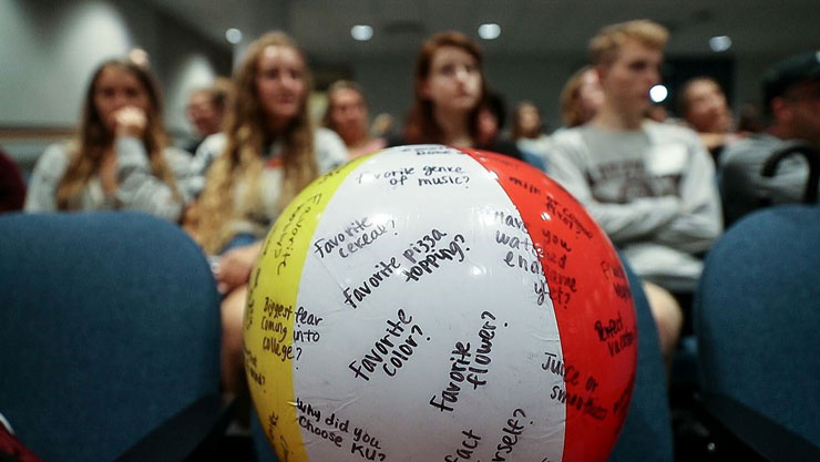 Closeup on a beach ball with questions on it, such as "what's your favorite color" and "why did you choose KU," with a group of students sitting in the background