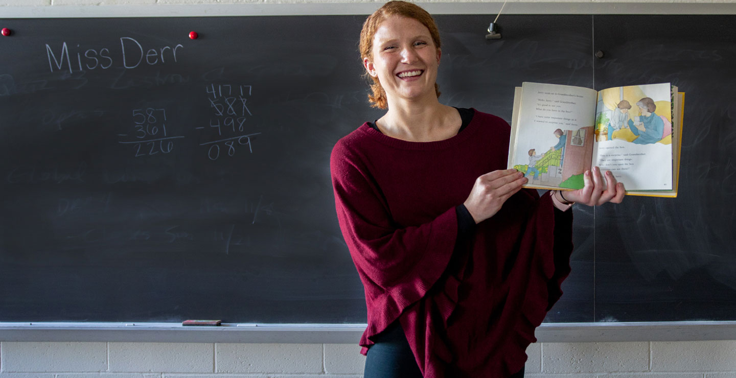 Photo of Rylee Derr standing in front of a classroom chalkboard holding an elementary picture book open.