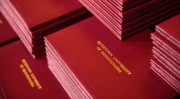Image zoomed in on multiple stacks of red diploma covers with "Kutztown University of Pennsylvania" imprinted in gold, capital letters. 