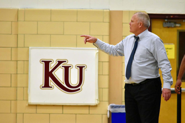Kutztown University's men's basketball head coach, Bernie Driscoll, coaches from sideline. KU logo banner (Maroon letters, "K" and "U" outlined in gold on white background) hangs on the wall in the background. 