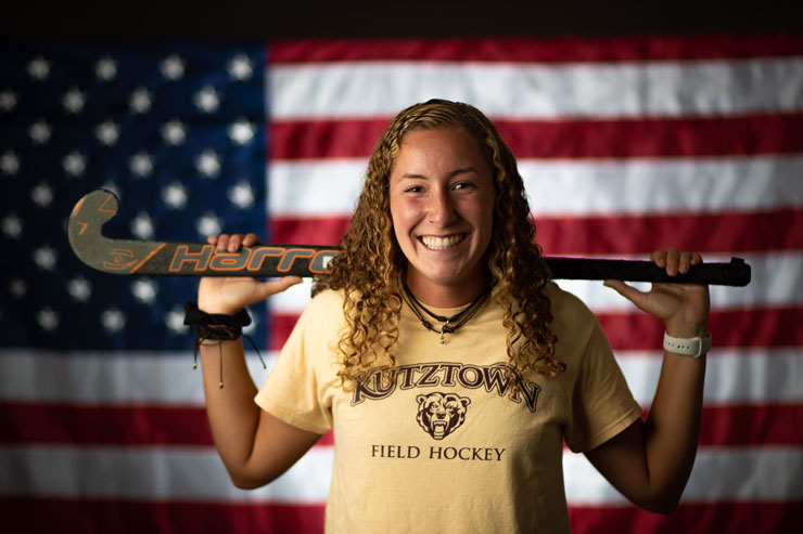 Flag of United States of America serves as backdrop in image of KU Field Hockey player, Erika Barbera, wearing yellow T-shirt with maroon type KUTZTOWN FIELD HOCKEY with the face/head of athletic bear in the middle, as she holds field hockey stick across shoulders behind her head. 