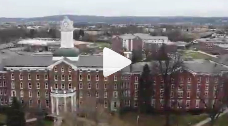 Click for Instagram Video - white play arrow centered over aerial image of  Old Main, looking towards south campus. 