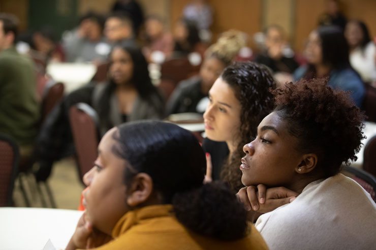 Photo focusing on three female students sitting at a table during a Black History Month Celebration. Other people in the background out of focus.