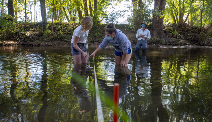 Three environmental science students conduct research while wading in river.