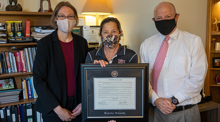 Coach Judy Lawes (center) holds Resolution awarded by the Kutztown University Council of Trustees and President, Dr. Kenneth S. Hawkinson. Director of athletics, Renee Hellert is on the left, and vice president for University Relations, Matt Santos is on the right. 
