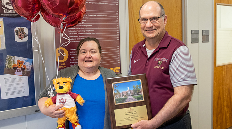 Photo of Letitia Manwiller (left), Employee of the Month for March 2021, standing with President Hawkinson (right). Tish is holding three maroon KU balloons, toy Avalanche, and EOM plaque.