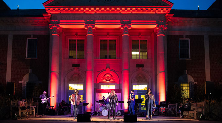Kutztown University's Rock Ensemble I performs in front of a red and purple illuminated Shaeffer Auditorium during a socially distanced nighttime music event.