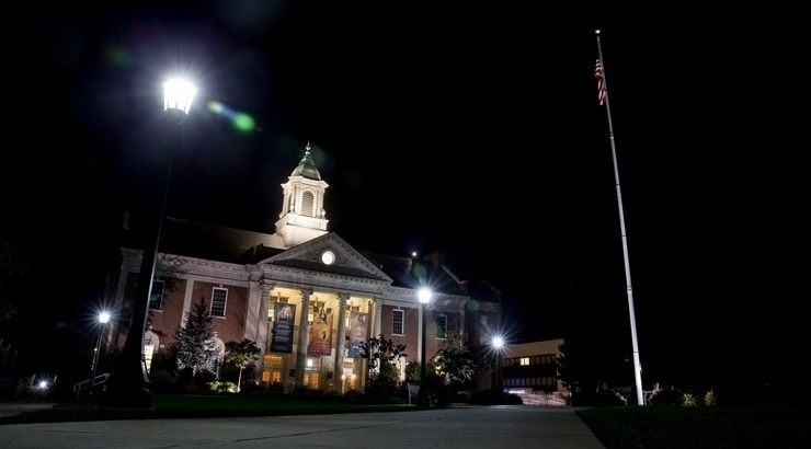 Nighttime image of Schaeffer Auditorium. Sidewalk lamppost to the left lights the path and an illuminated flag pole is on the opposite side of walk way.