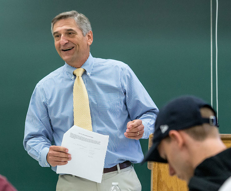 Professor David Wagaman standing in front of a chalkboard as he reviews content of stapled sheets of paper with the class. 