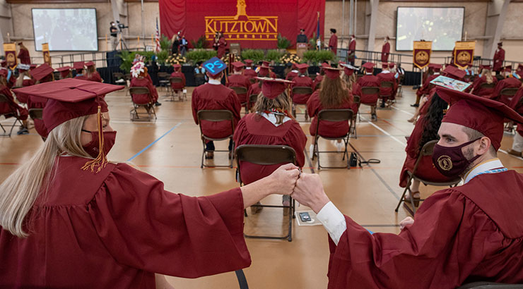 Looking towards the stage, two Kutztown University graduates fist bump while seated socially distanced at the rear of the auditorium during commencement. 