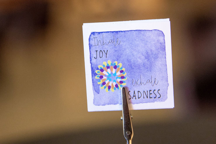 Picture of hand decorated card promoting health and wellness. Card features a purple stamped square with a yellow, purple and blue mandala with the words "Inhale Joy" at the top left corner and the words "Exhale Sadness" at the bottom right corner.