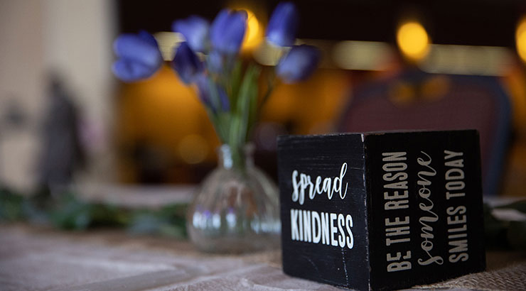Photo of a desktop. Cube has "spread kindness" on one side, and "be the reason someone smiles today" on another side. Purple flowers in clear glass vase in the background.
