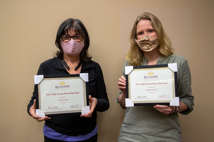 Drs. Watson (left) and Oakley (right), wearing ppe masks, pose for photo op holding  their Undergraduate Research and Creativity Mentorship awards. 