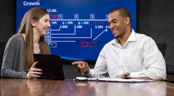 Two students sit at a desk reviewing content on a notepad. A blue data chart is on the screen behind them. 