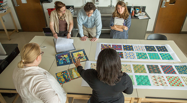 Members of Kutztown University's Communication Design Program gather around a drafting table to review multiple pieces of digital artwork spread out on the table.