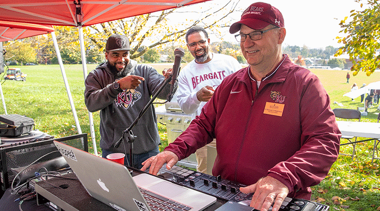 Photo of President Hawkinson DJ at homecoming. Two men watching and cheering for him.