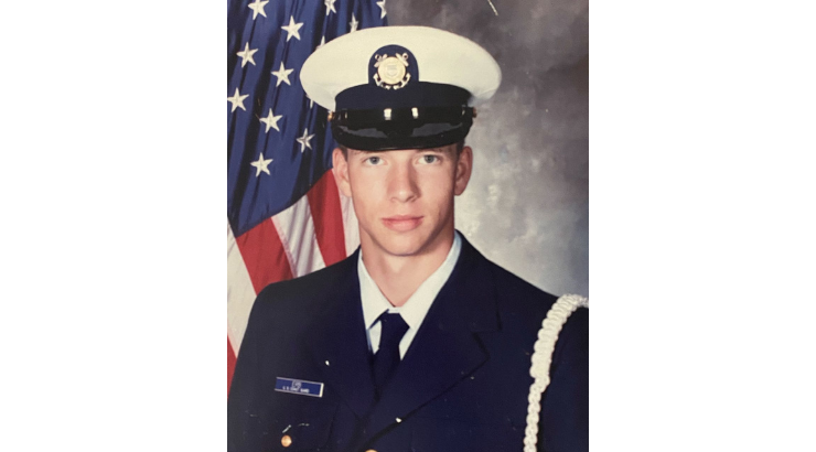 Formal studio portrait of a person in a military dress uniform, standing in front of a United States flag backdrop.