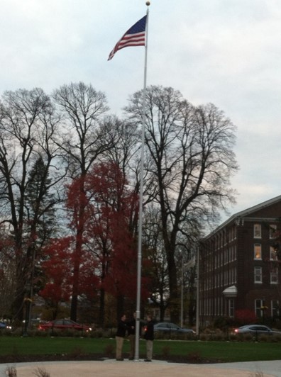 an image of the flag on campus. Main street and the bell plaza in the background with fall trees