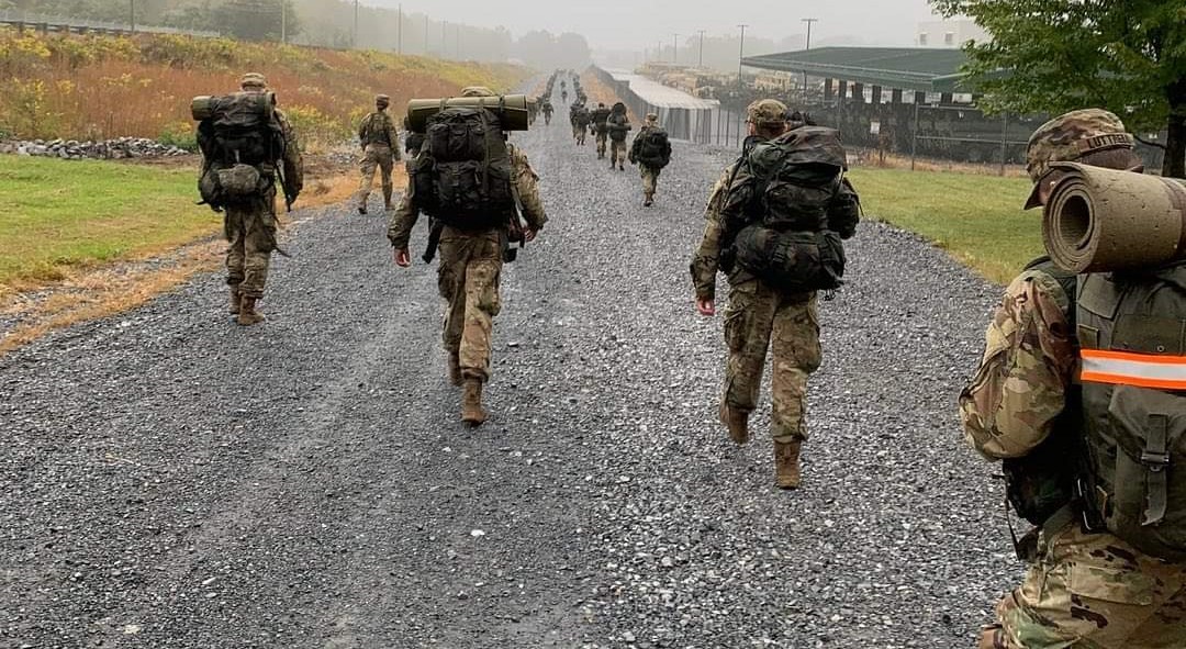 ROTC students marching with their rucksacks on a road. A quote is in the picture that reads, "we are what we repeatedly do. Excellence, then, is not an act, but a habit." Aristotle