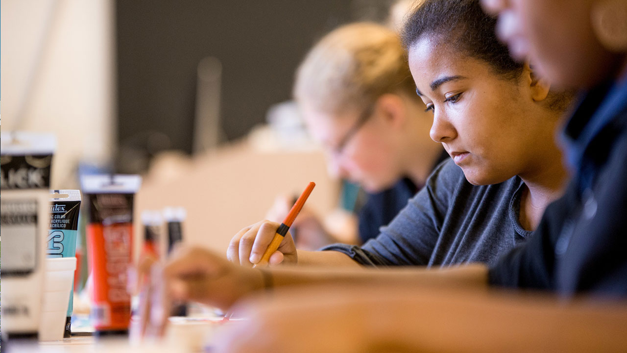 Three art students painting at a table with tubes of paint in front of them