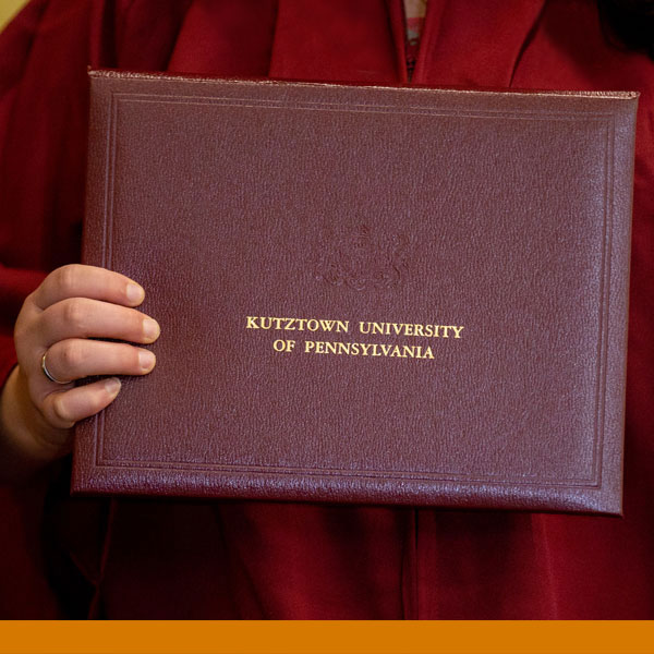 image of a hand holding a ku embossed diploma against the traditional maroon robe worn by graduates