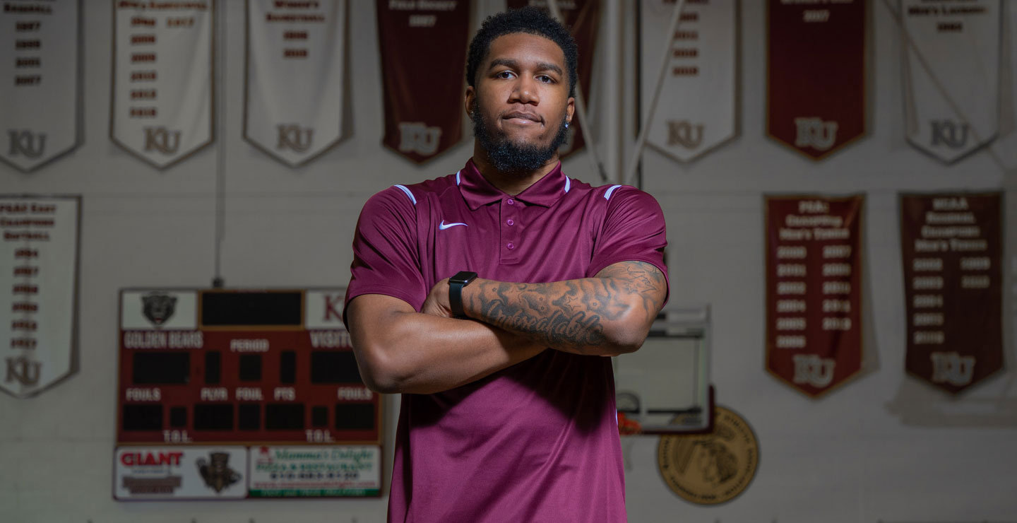 Male student with arms crossed, looking at camera, standing in front of championship banners.