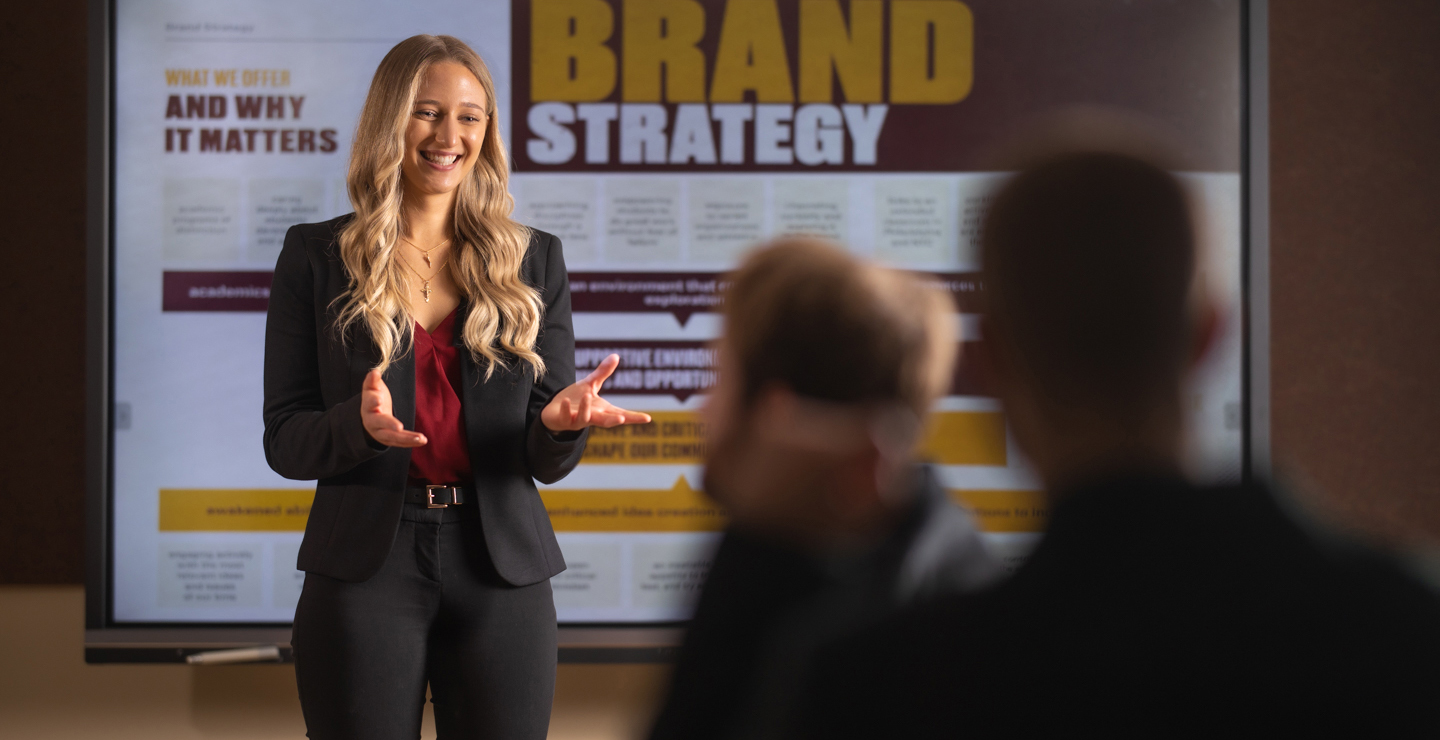 Female student with marketing display behind her, presenting to audience in foreground.