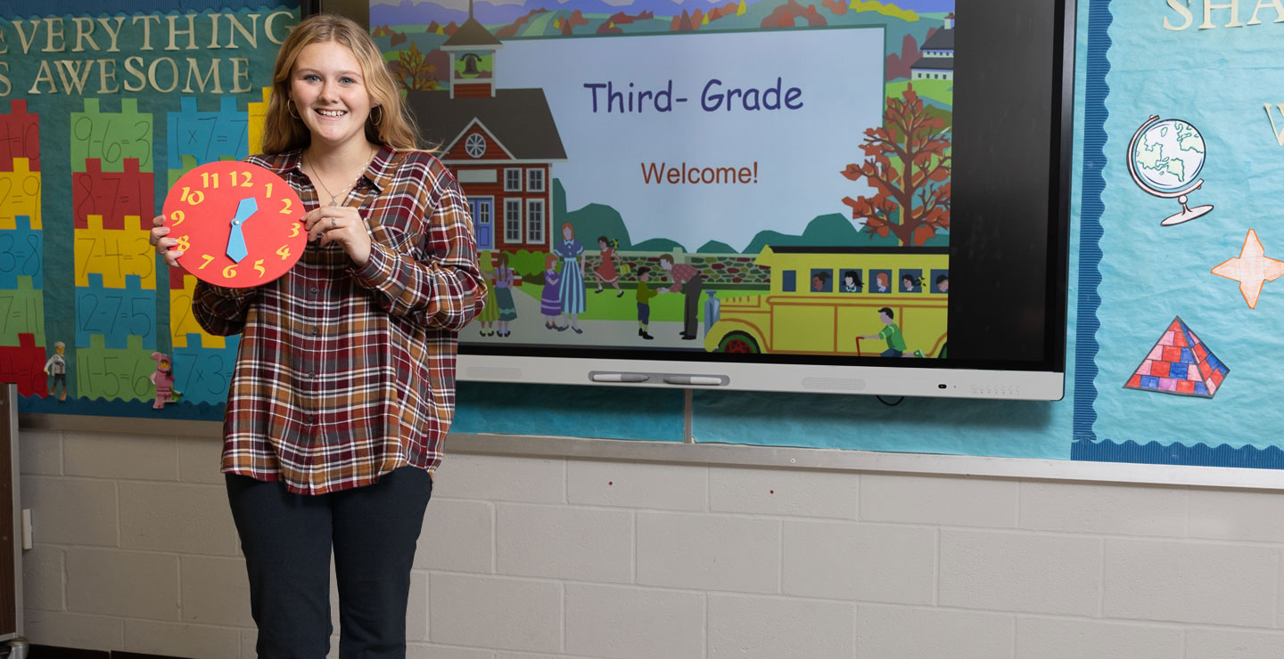 image of a female student standing in front of a digital school board holding up a clock