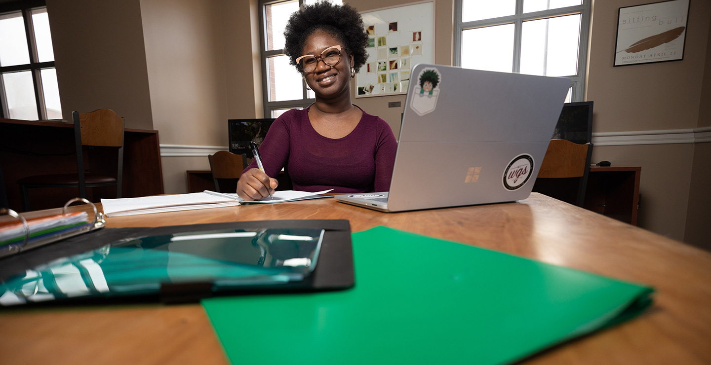 Professional Writing Major Eva writes on a pad, with a laptop to the side
