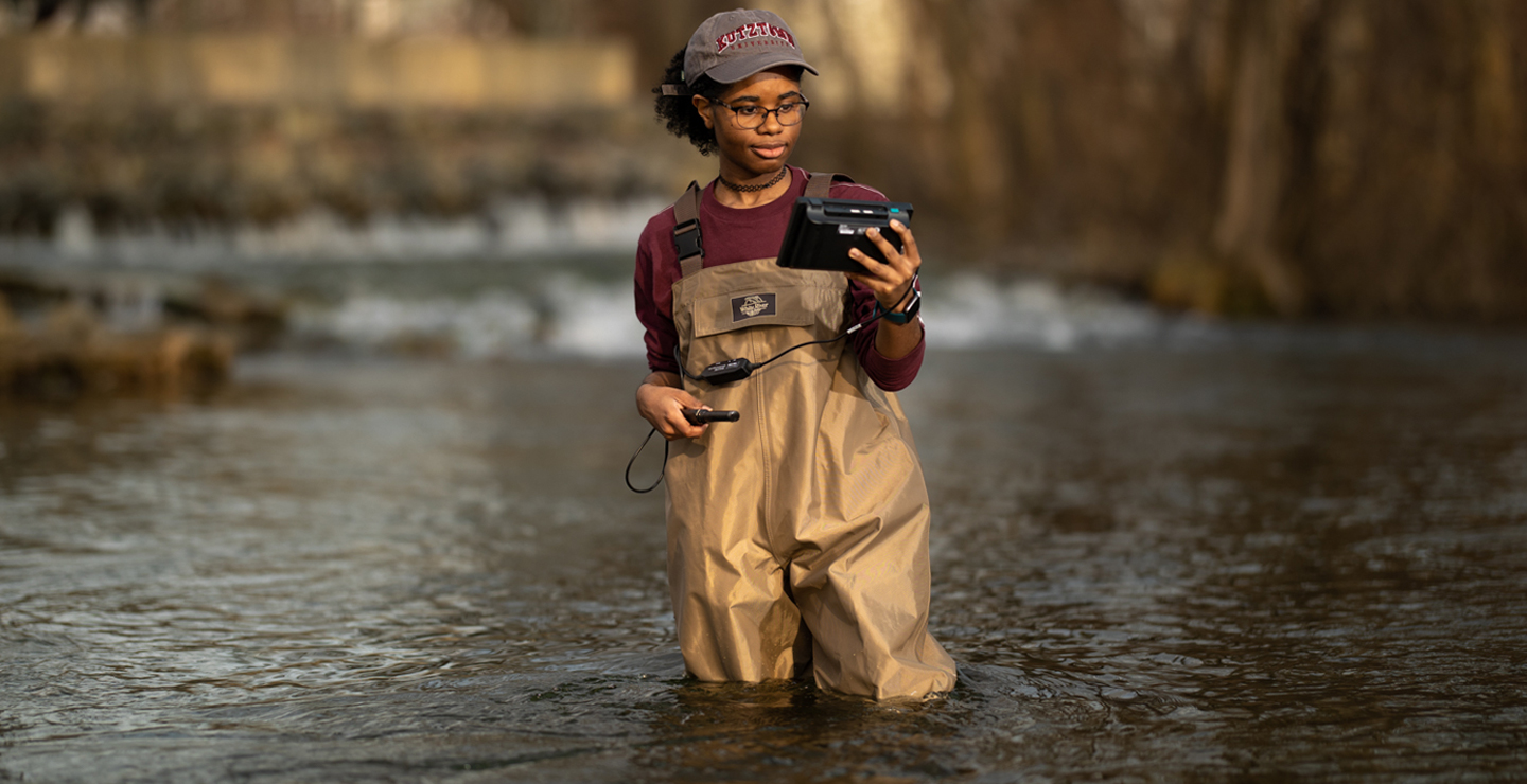 Female student on location looking at readings on a divice while standing in a river.