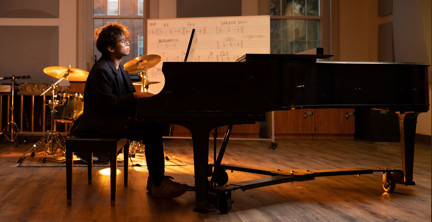 Male student sits at a grand piano, with other musical instruments and musical writing on a whiteboard behind him. 