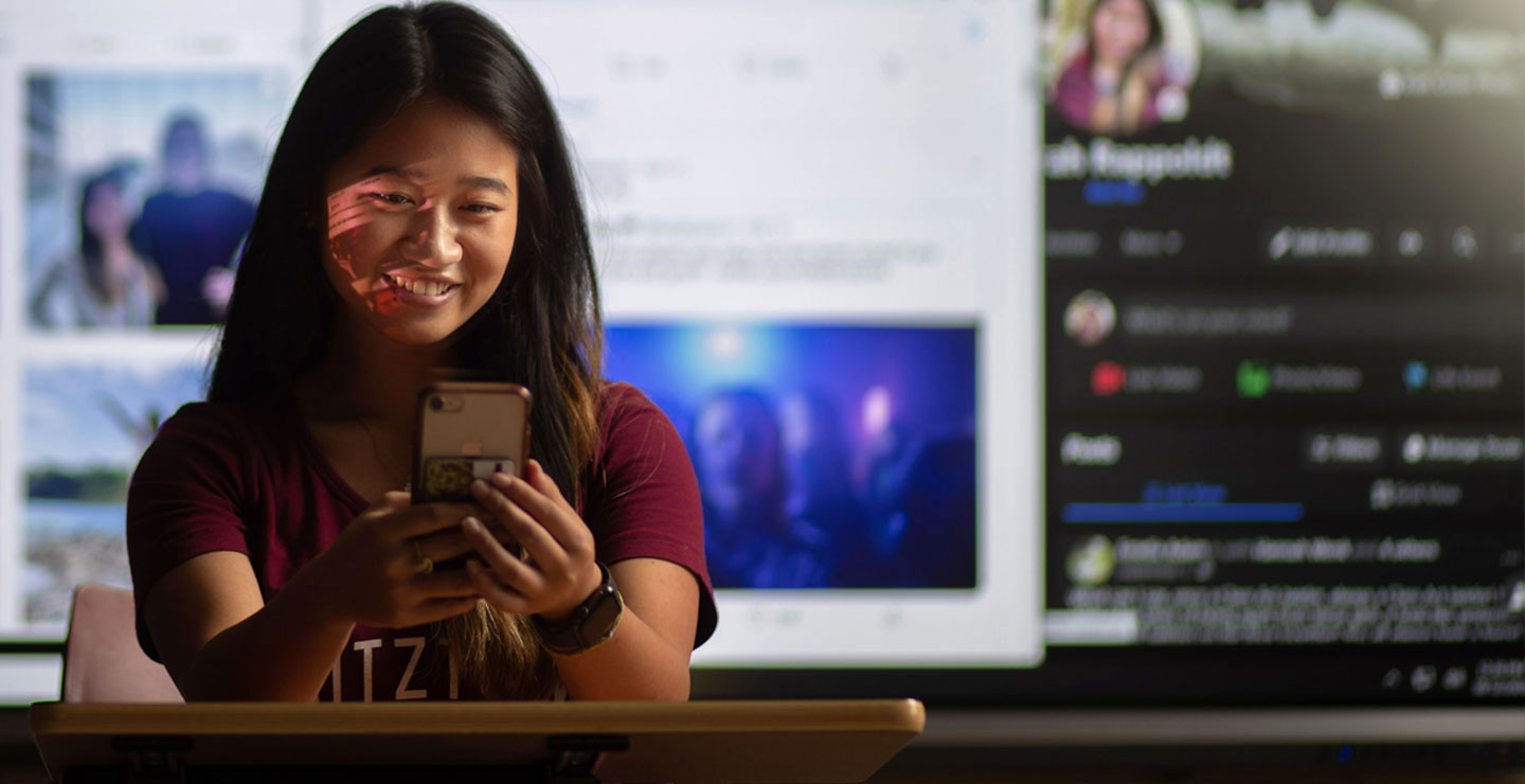 Female student working on her mobile device, standing in front of various social media displays.
