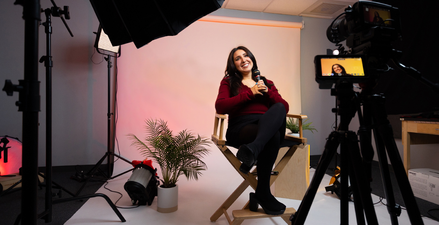Female communication studies major sitting in a directors chair, holding a microphone. In the foreground is a camera's viewfinder, that shows another angle of the student.