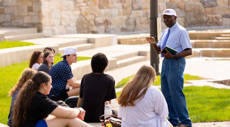 Male faculty member lectures to students gathered on the alumni plaza on a spring day.