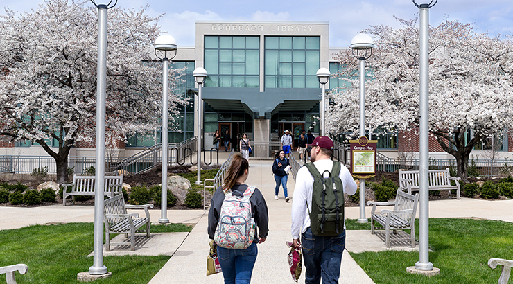 Students on path walking about to enter Rohrbach Library surrounded by green grass and two trees with white flowers