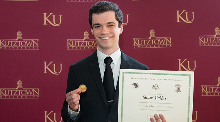 Issac Reiter holding the Chambliss Academic Achievement Gold Medal certificate in front of wallpaper with the Kutztown University logo 