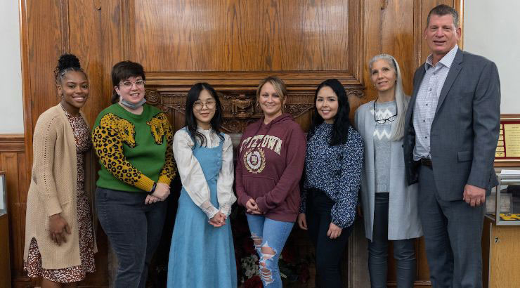 A group of seven diverse people standing in front of a wood panelled wall (Old Main Concourse)