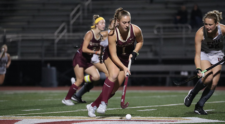 Female field hockey player hitting a ball on the field, surrounded by other players