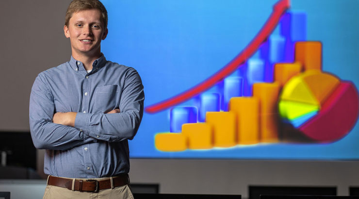 Business student with arms crossed stands in front of projection of graphs.