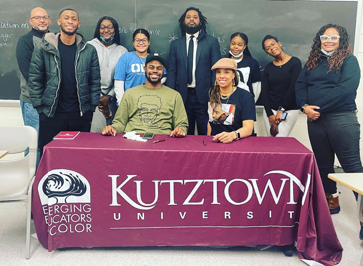 Members of emerging Educators of Color smiling and standing or sitting as a group behind a table with a Kutztown University tablecloth 