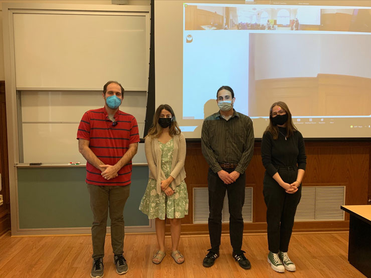 Three students and professor wearing masks and standing in front of a screen with a zoom class open