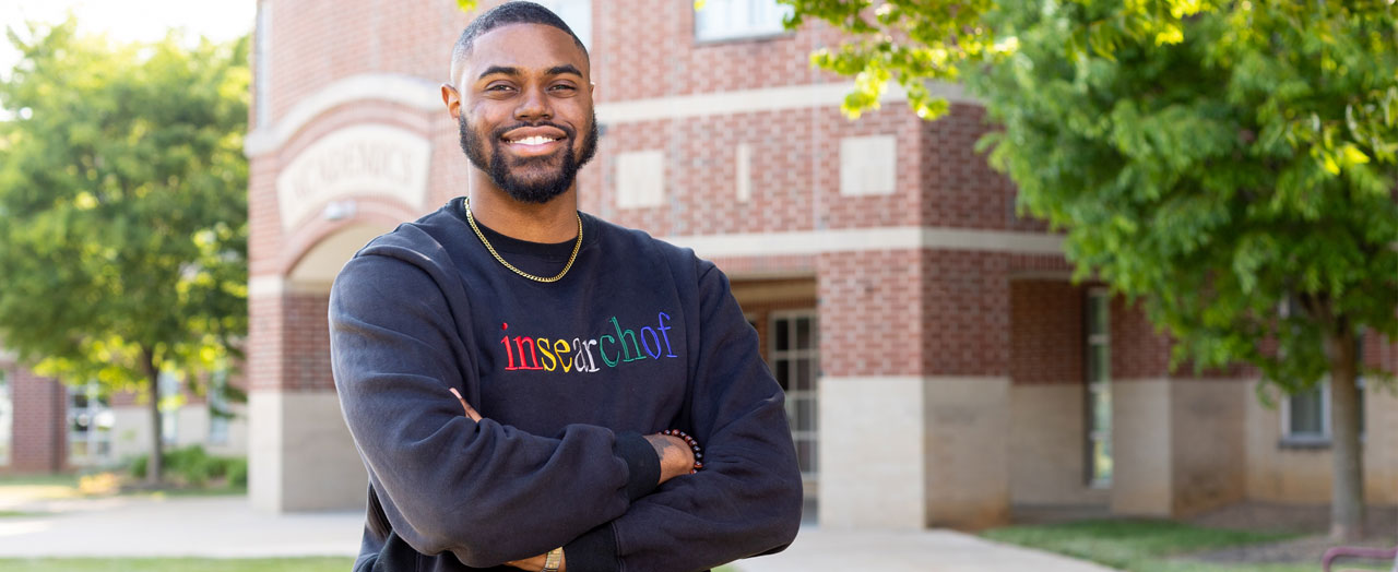 Akello Mosby stands with arms folded in front of a brick building and green trees. His blue sweatshirt reads, in colorful letters, "in search of"