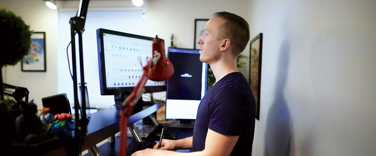 Griffin Macaulay working at a standing desk with two computer monitors and a red desk lamp. 