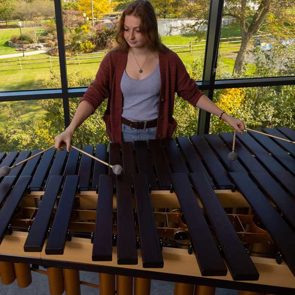 Female commercial music major standing infront of a window showcasing the landscape of campus also playing a marimba