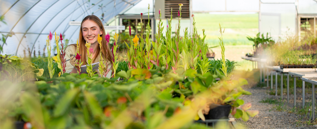Female Kutztown University Regenerative Organic Agriculture student smiles amidst an array of plants in the Stump-Seiger Botantical Research Center at Kutztown University.