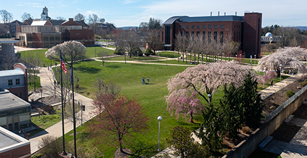 North Campus overlooking the quad on a bright, sunny day, facing toward Boehm and Schaeffer Auditorium. Green grass and colorful trees adorn campus in spring-time. Students are walking around campus, as the American and Pennsylvania flags fly atop flagpoles in the foreground near the McFarland Student Union.