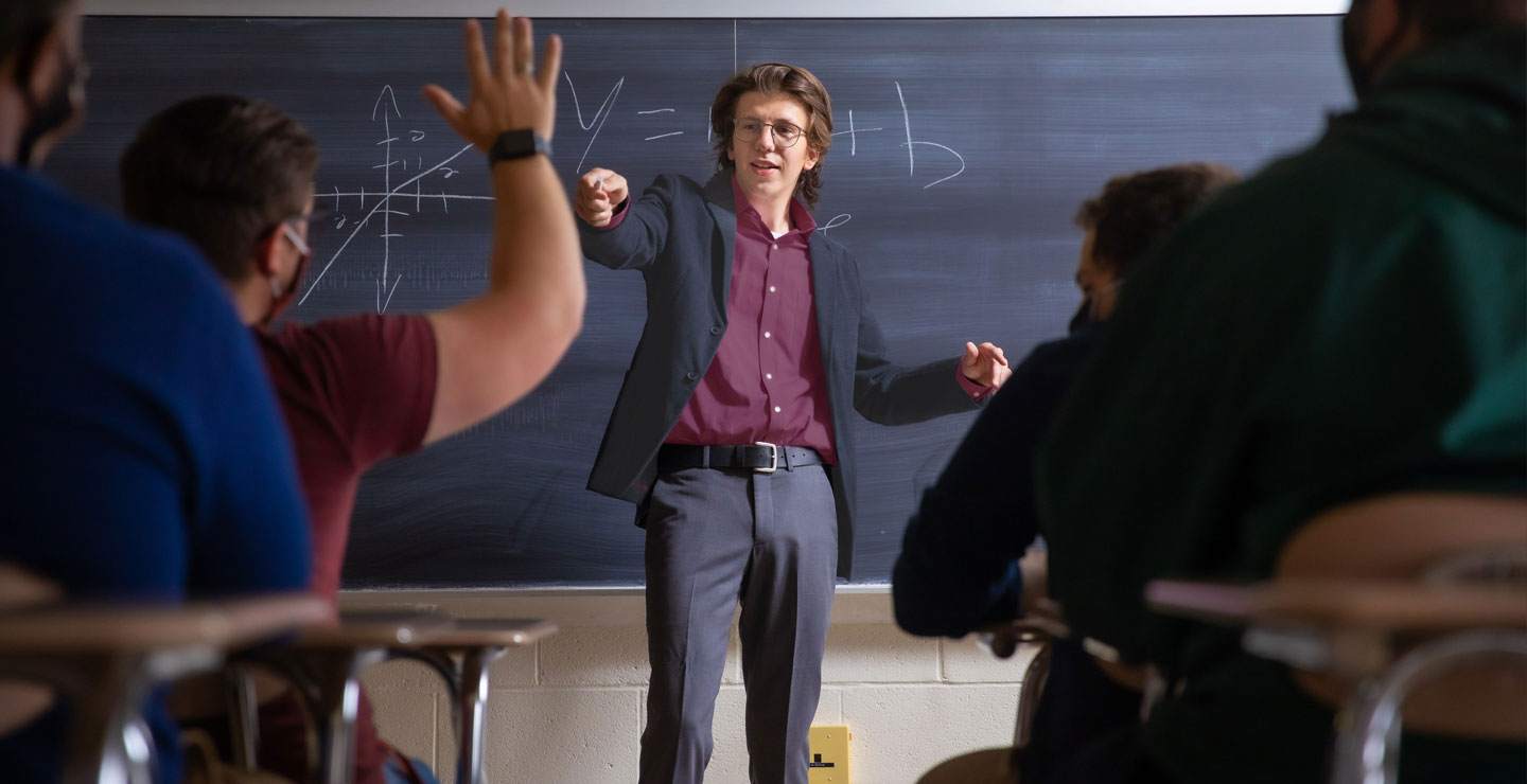 Male teacher in classroom pointing at student with hand raised.