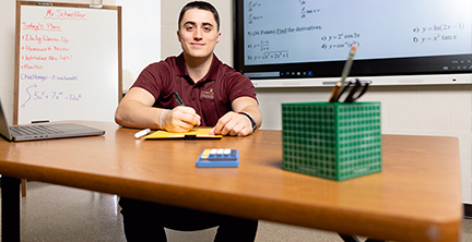 Male math education major holding a pen above a pad.  In the foreground is a blue calculator, green box with supplies and a laptop computer.  In the backgrround are math formulas and questions, as well as a white board that reads "Mr Shcaeffer, Today's plan: Daily Warm-up, homework review, introduce new topic, practice, challenge evaluate."